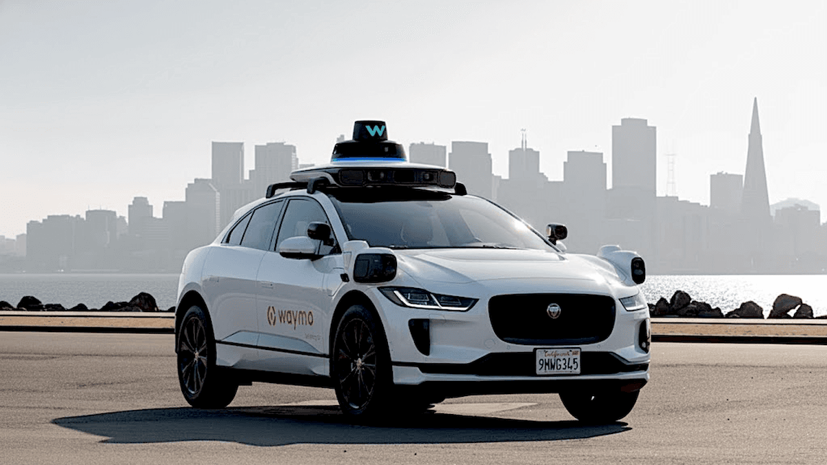 White Waymo vehicle near water, city skyline visible; displays autonomous service for urban freeways.