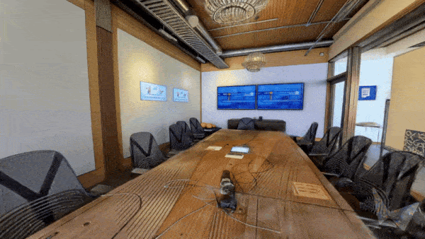 GIF showing a 360° walkthrough of a conference room with a wooden table, high-back chairs, wall screens, and ceiling lights.