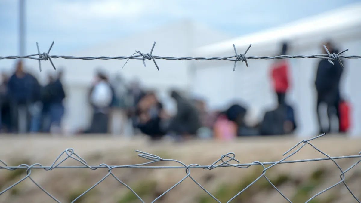 Detained migrants wait and talk behind a chain link fence.